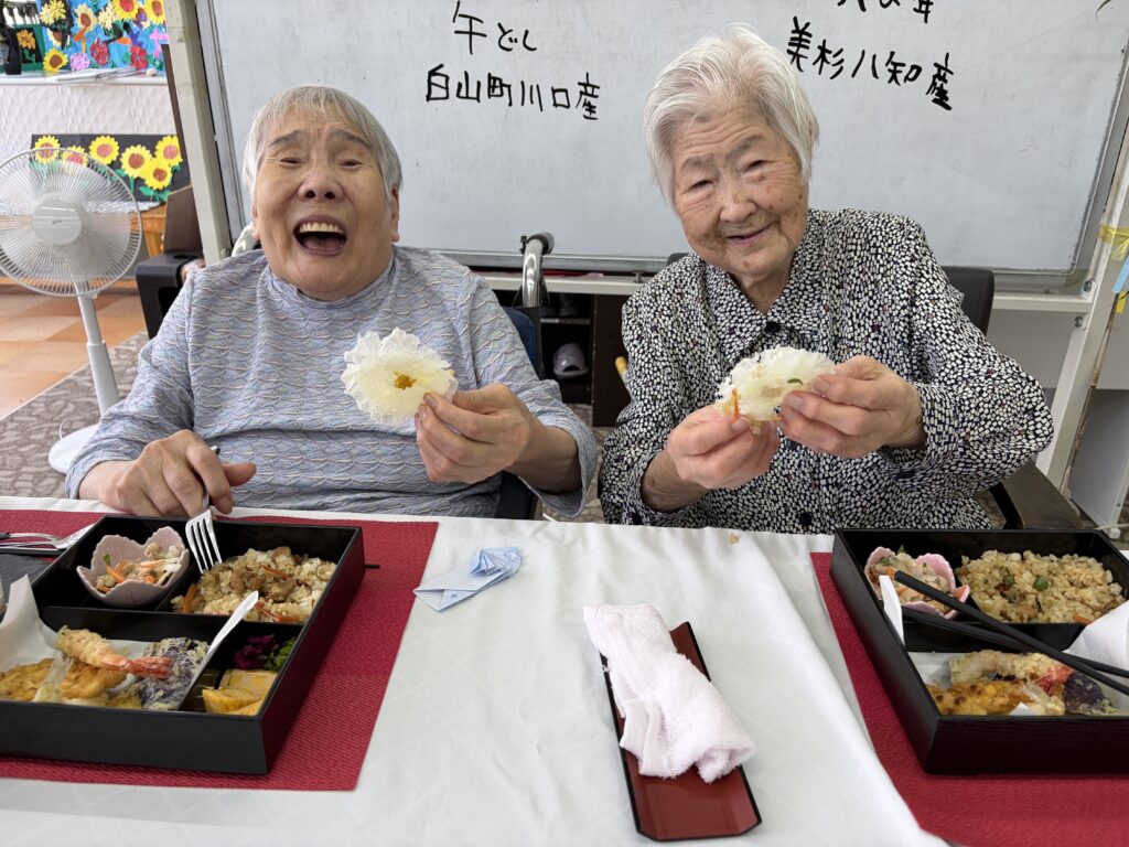 お花の飾りを手に、笑顔を見せるご利用者様おふたりとお祝いのお弁当
